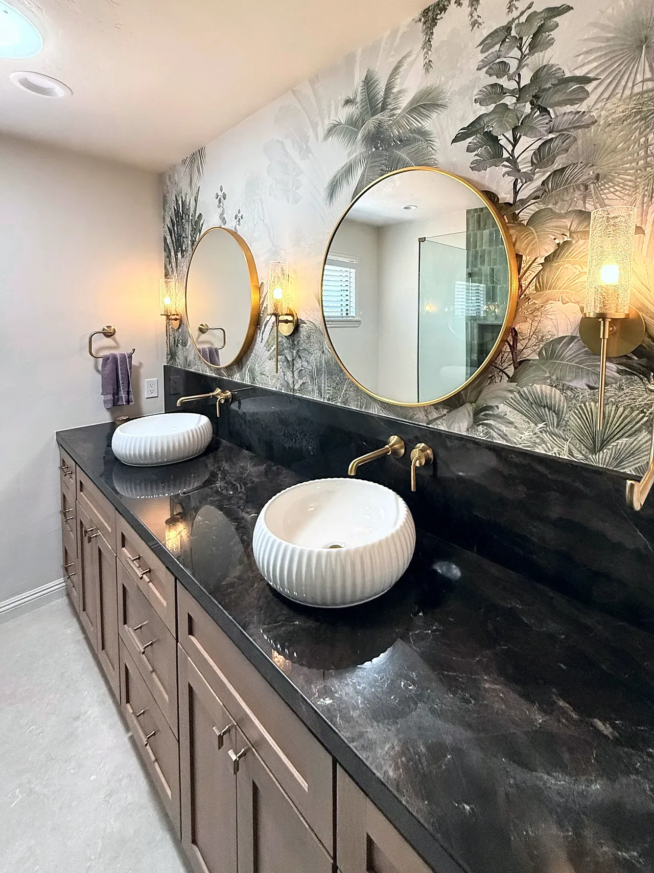 The finished vanity wall: round brass mirrors, vessel sinks, a dark stone top and botanical wallpaper