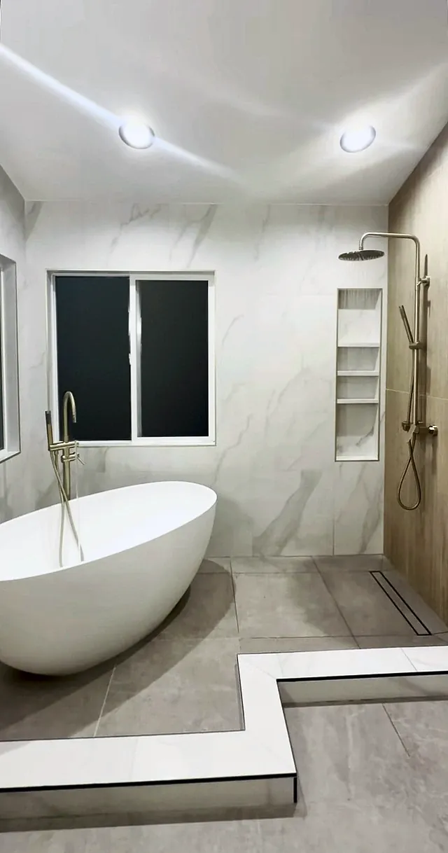Freestanding tub with a floor-mounted brushed gold filler next to a walk-in shower with a tall niche and a linear drain