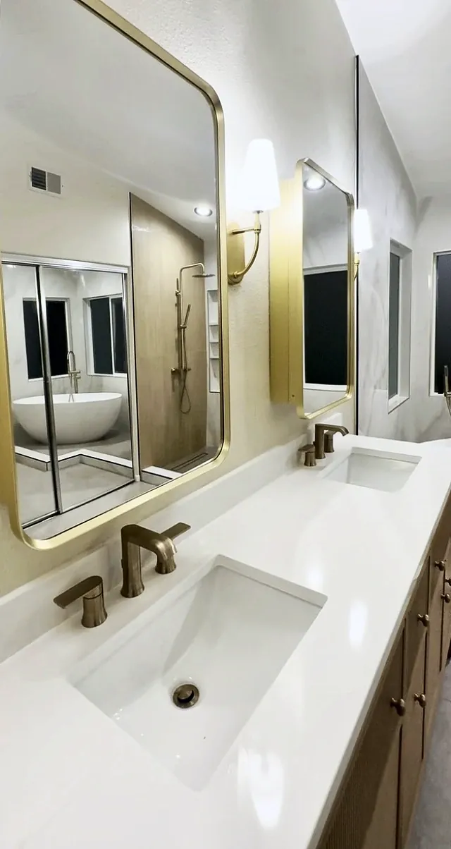 White vanity top with an undermount sink and a brushed gold faucet, with a brass mirror reflecting the tub and shower