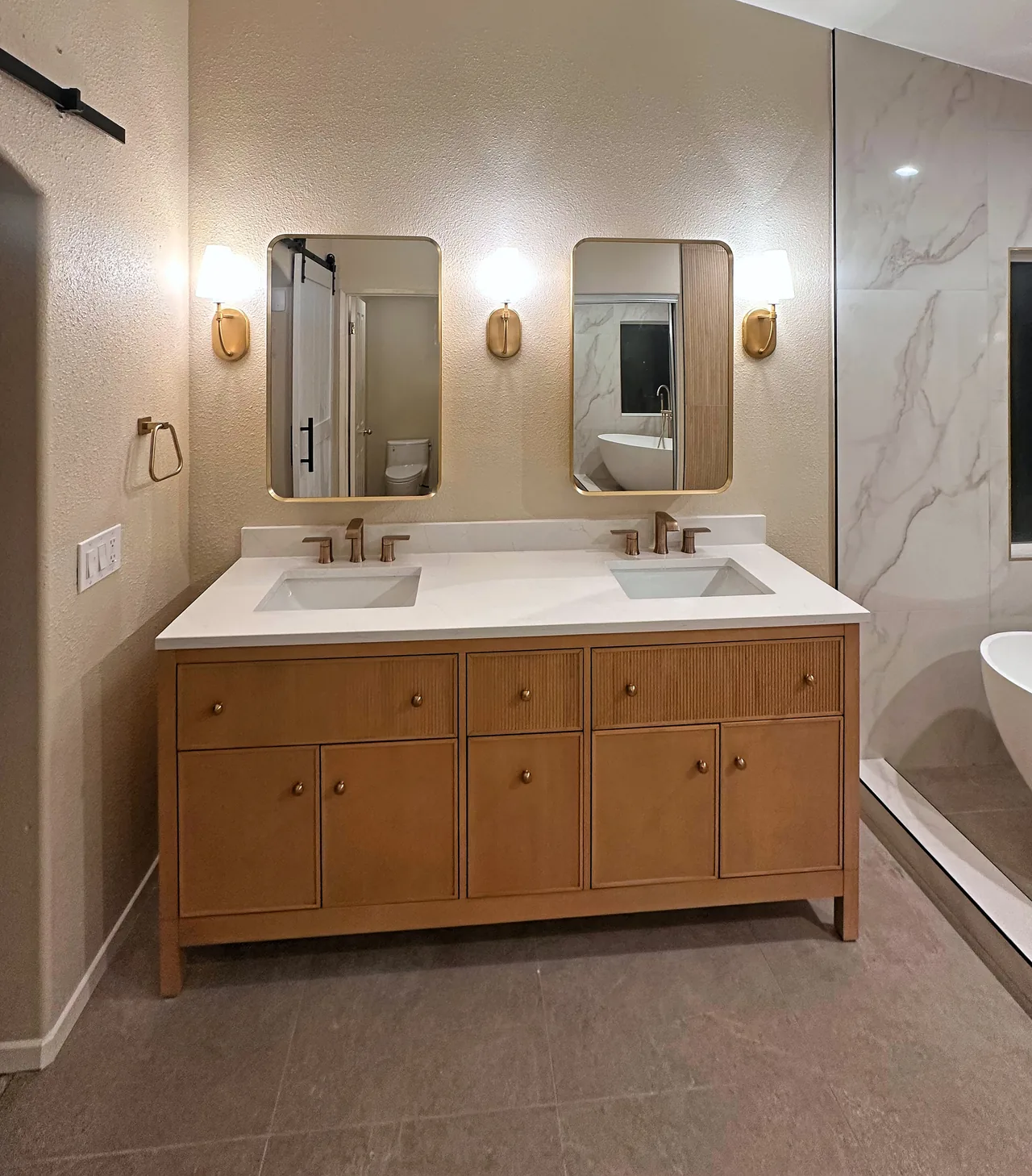 Light oak double vanity with fluted drawer fronts, a white top, two brass mirrors and three sconces, beside the glass of the walk-in shower
