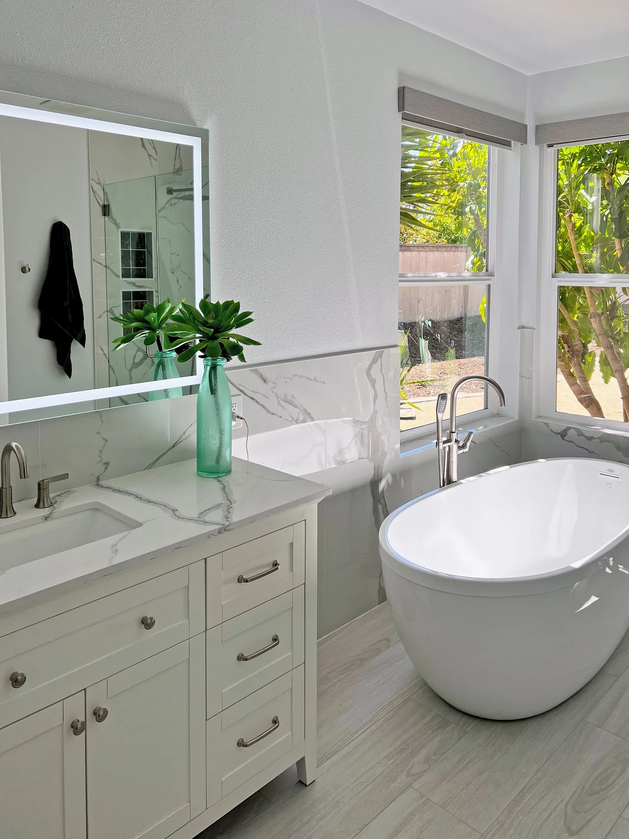 White shaker double vanity under a lit mirror, with a freestanding tub by the corner windows