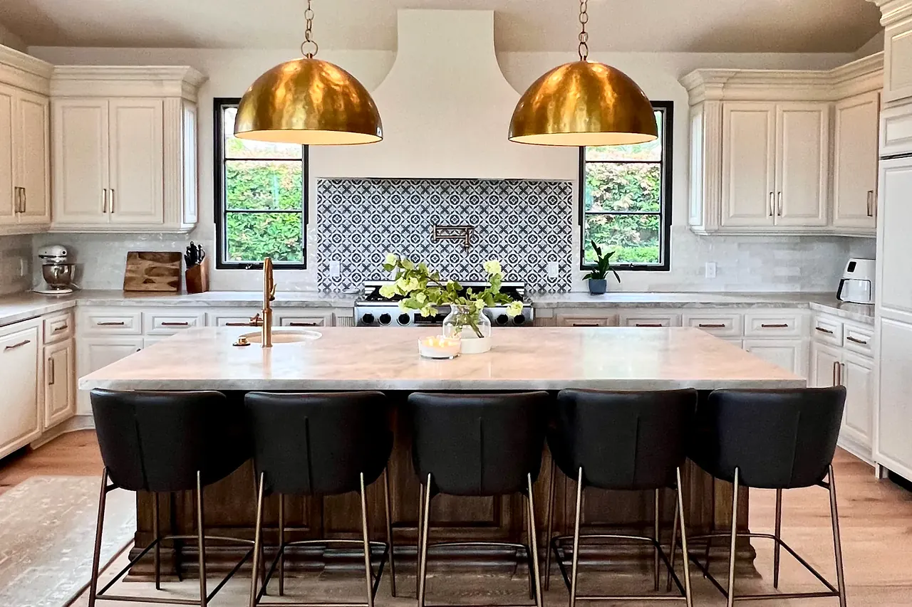 Finished kitchen with two hammered brass dome pendants over a large island, white raised-panel cabinets and a patterned tile backsplash between black-framed windows