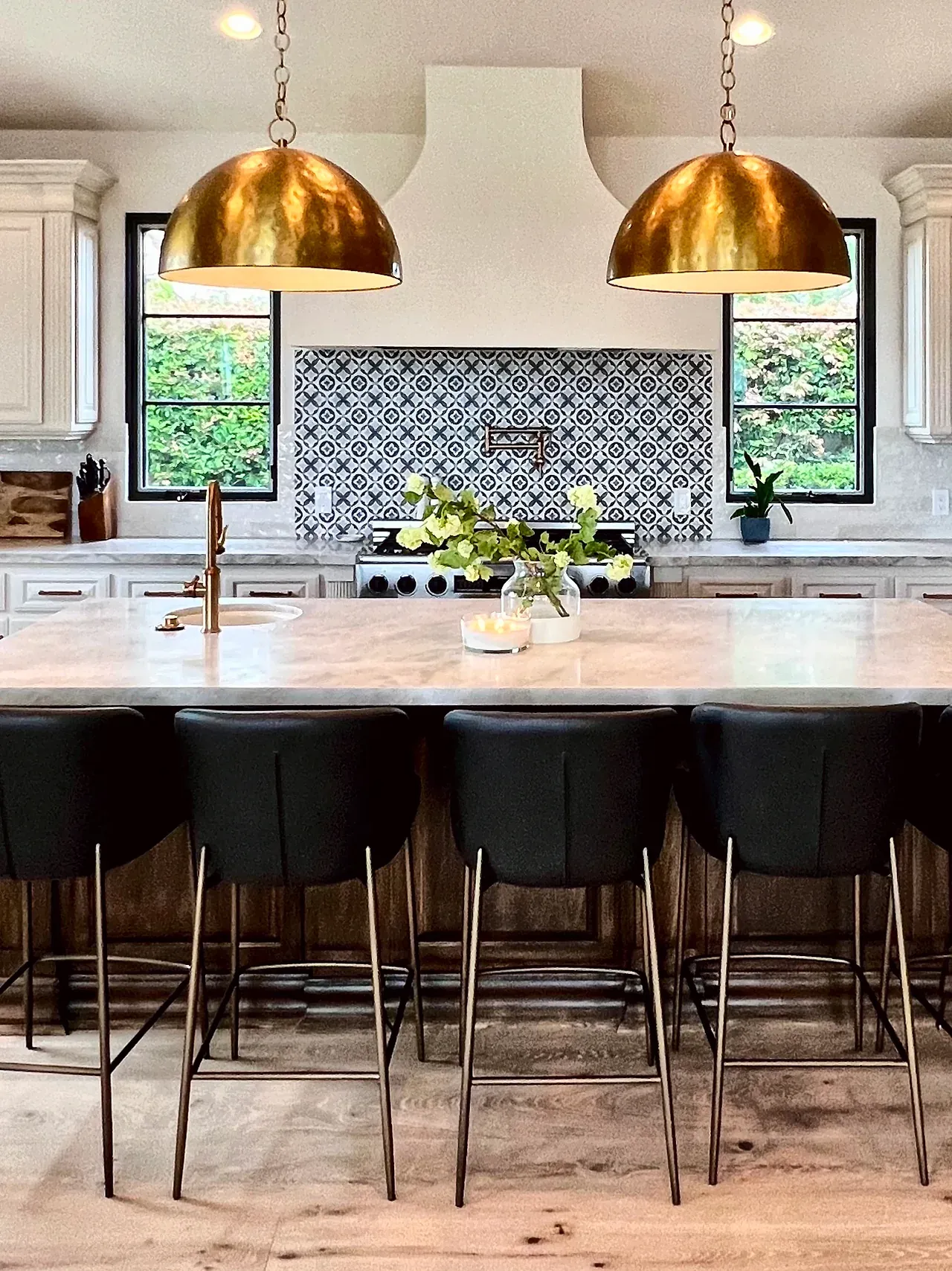 The same Santaluz kitchen after the remodel, with cream cabinets, a smooth white range hood, a patterned tile backsplash, black window frames, brass pendants, light wood floors and a new stone island top
