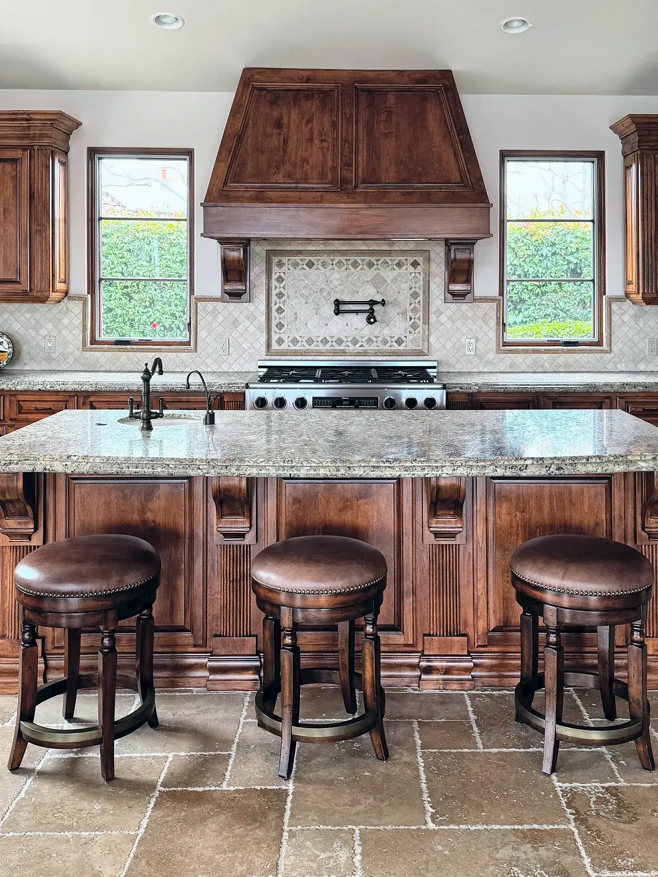 Santaluz kitchen before the remodel, with dark stained cabinets, a wood range hood, tumbled stone floors and a granite island with three counter stools